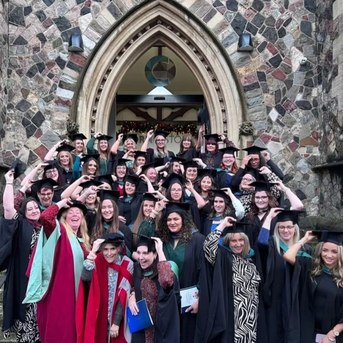 Around forty Early Years graduands in academic robes stand in front of a Victorian Gothic arched stone doorway, smile, and hold their mortarboards.