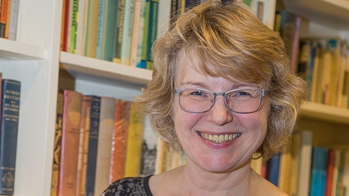 Yn sefyll o flaen cwpwrdd llyfrau, mae’r Athro Bettina Schmidt yn gwenu tuag at y camera wrth ddal dau o’u gweithiau ei hun: “Astudiaeth o Brofiad Crefyddol – Dulliau a Methodolegau”; hefyd, “Ysbrydion a Llesmair ym Mrasil”.