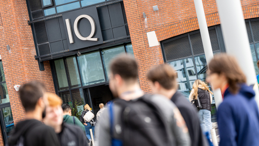Students chat outside the entrance to a modern brick building on which the silver letters IQ stand out from black panels above the sliding glass doors. 