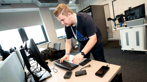 A young man uses a keyboard; a bag of tools lies open to his right; two phones and an external hard drive are on the table to his left.