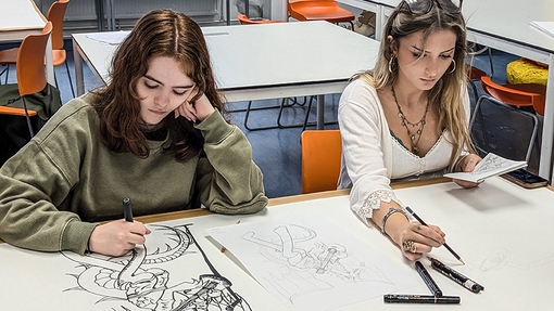 Two college students looking down at their illustration work, sitting at a large white table in a workshop.