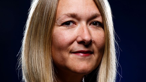 A smiling lady dressed in a patterned top, looking at the camera, against a black backdrop.