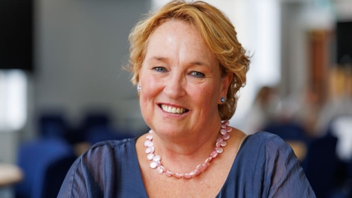 A smiling lady in a blue top, wearing a necklace, sitting in a conference-style environment.