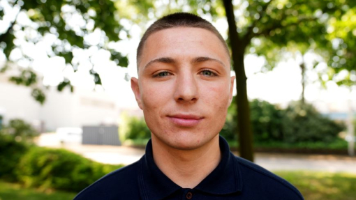 A student in a dark polo top standing outside with trees in the background.