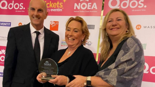 A man and two women dressed in smart evening dress, celebrating and holding an award against a branded background.