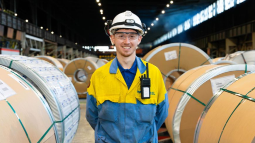 A degree apprentice student dressed in high vis, protective gear standing between rolls of manufactured steel.