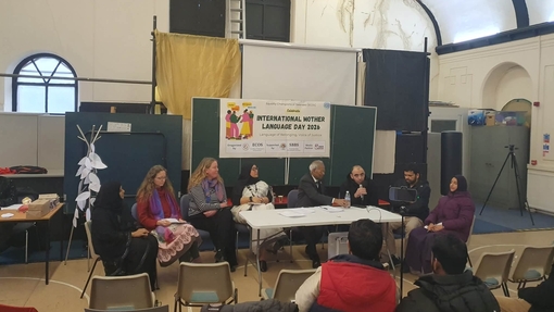 Eight people sitting as a panel behind a table at the front of the hall with a International Mother Language Day banner hung up behind with a 
