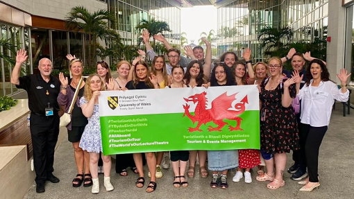 A large group of lecturers and students standing outside holding a banner with the Welsh flag.