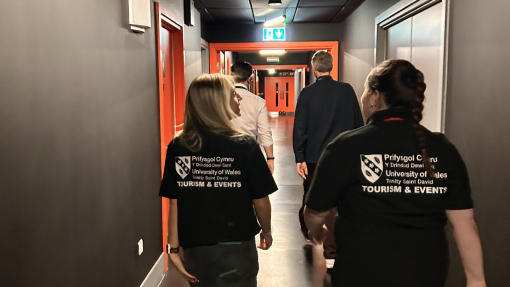 Three students with their backs to the camera walking down a dimly lit corridor in an arena style building.