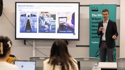 A man dressed in a suit delivering a talk in front of a white screen in front of an audience in a lecture room.