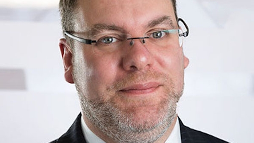 A cropped portrait image of a man wearing glasses and a suit and tie.