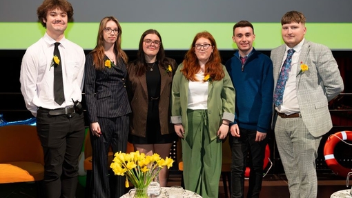 A group of students standing in front of a large screen in an arena, with a table with daffodils in a glass jar in the centre.