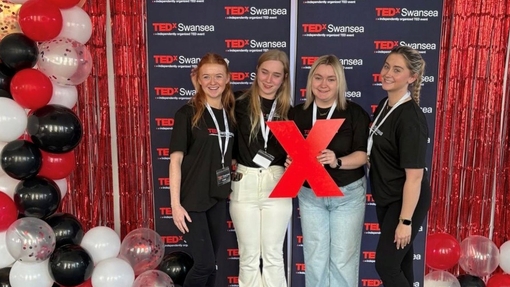 Four students standing between a red, white and black balloon arch in front of a red and black branded background.