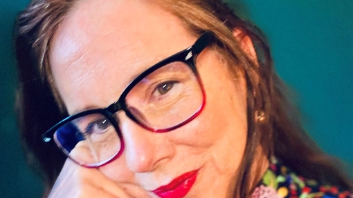 A woman wearing glasses with her right hand raised to her face and wearing a multi-coloured top.