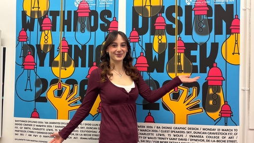 A girls holding up her arms as if she's displaying the wall behind her. On the wall behind her, there is a graphic that reads 'Design Week 2026'