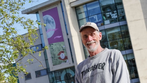 A man in a baseball hat and grey sweatshirt smiling and standing outside a glass-fronted building with a giant canvas at the top.