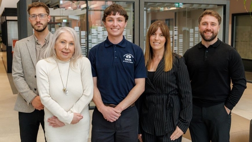 A group of five degree apprentices, smiling and standing proudly against a glass fronted background.