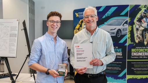 Two men standing in front of an exhibition area, one is holding a trophy, the other a certificate.