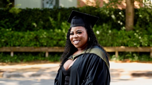 A graduate standing, turned to the side, smiling and proud, dressed in her cap and gown.