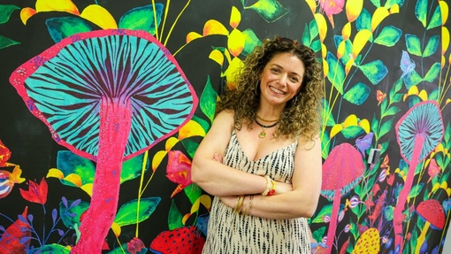 A smiling graduate standing with her arms folded in front of a vibrant mural.