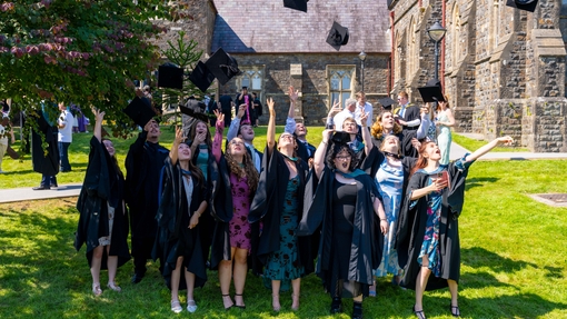 Group of students in graduation gowns standign on grass bank in front of Carmarthen's old college building throwing hats in the air