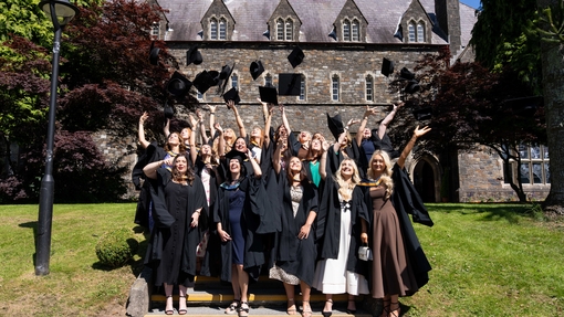 Group of students wearing graduation gowns standing in front of the old college building throwing their hats in teh air
