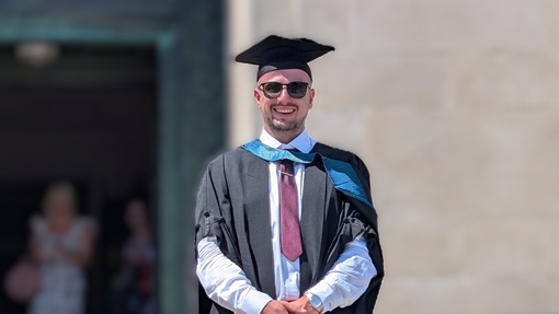 A smiling and proud graduate standing in his cap and gown on the steps in front of a stone-walled building..