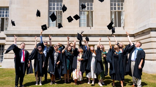 A large group of graduates dressed in caps and gowns celebrating by throwing their hats into the air, standing outside a stone-coloured building with large windows in the background.