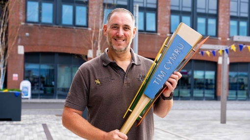 A man in a brown T-Shirt holding a wooden replica torch, standing in front of a red brick building.