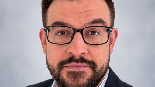 A headshot of a man wearing glasses, wearing a suit coat and shirt.
