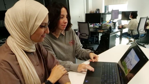 Two University staff sat at a white desk looking at technology.