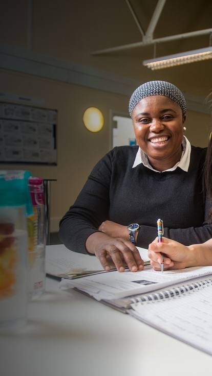 Two people seated at a table in a bright study space, working together on a notebook and smiling toward the camera, with pens, papers, and a water bottle visible in the foreground.