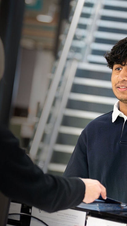 Person at a reception desk handing over a card to a visitor in a modern building, with a staircase visible in the background.
