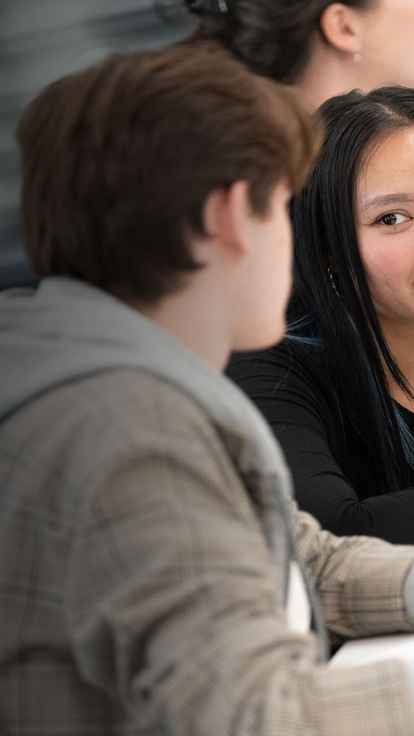 Two students seated at a table, looking at each other while discussing work, with a tablet and papers on the desk and others blurred in the background.
