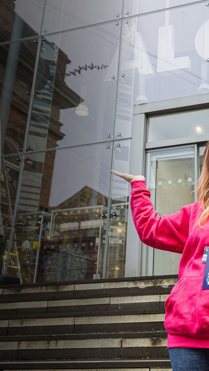 Person in a pink UWTSD hoodie standing outside the university entrance between UWTSD flags.