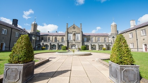Picture of outside Lampeter Campus, grey brick building with grey flagstone walkway with a water fountain in the centre and two green bushes in grey square plant pots on a sunny day