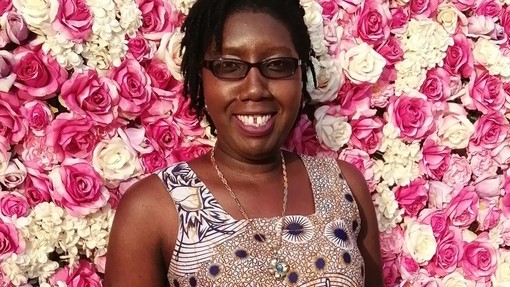 Tendai Nzirawa yn sefyll yn gwenu o flaen wal o rosod pinc a gwyn.