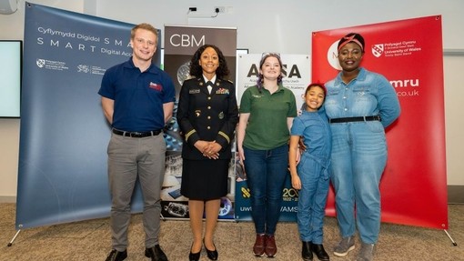 Flight Lieutenant Jack DeSchoolmeester, Major Rosette Clarke-Morton, UWTSD Electrical / Electronic Engineering graduate Carys Williams, a pupil from Cefn Saeson Comprehensive in Neath and Patricia Mawuli Porter OBE are standing in a classroom in front of four banners.
