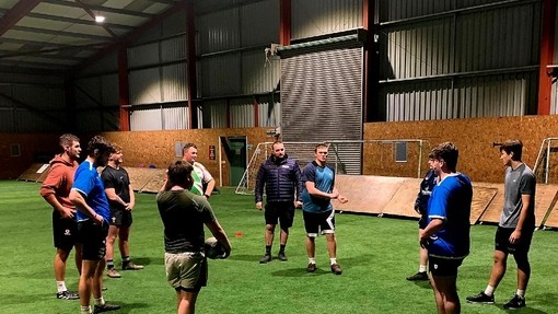 Ken Owens gyda thîm rygbi myfyrwyr ar gae dan do gyda llifoleuadau.