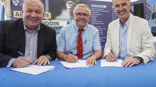 Tri dyn yn sefyll o amgylch bwrdd glas yn llofnodi dogfennau.