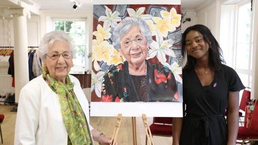 UWTSD BA Illustration student presenting her portrait of a lady who was affected by the Holocaust, genocide or identity-based persecution. The two females stand each side of the portrait smiling, the portrait looks exactly like the lady with flowers in the background of it.