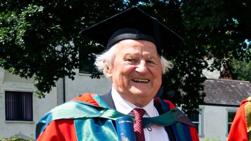 Dr Ned Thomas yn sefyll mewn cap a gŵn graddio ac yn gwenu.