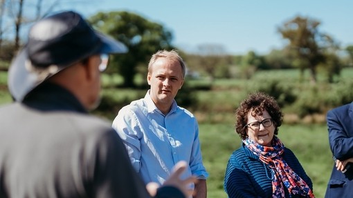 Three people standing outside surrounded by grass, trees and blue skies. A man in a hat is facing them and conversating.