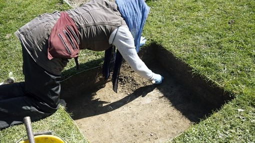 A person kneeled over carrying out excavation work, digging and cutting a square hole into the grass down to the brown earth.