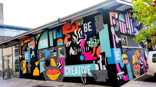Dwy ochr i adeilad unllawr â murlun lliwgar wedi’i baentio’n drwchus arno: mae’r geiriau ‘Be You, Be Creative’ i’w gweld ymhlith siapiau lliw.