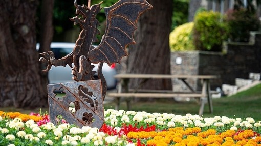 A metal sculpture of a Welsh dragon rears over a metal heraldic shield in a flowerbed filled with white and orange chrysanthemum.