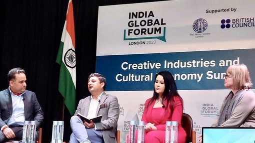 Gan afael llyfr ar agor ar ei lin, mae Dr Jaideep Gupte yn siarad wrth i dri phanelydd wrando; baner Indiaidd ac arwydd mawr yn dweud ‘Creative Industries and Cultural Economy Summit’ i’w gweld tu ôl iddynt.