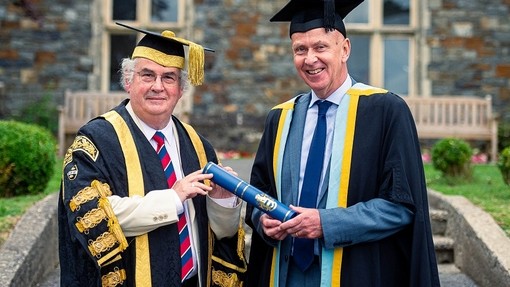 Yr Athro Medwin Hughes ac Ian Gwyn Hughes yn gwenu wrth iddynt gydio yn y sgrôl las sy’n symboleiddio’r Gymrodoriaeth gyda’i arfbais gilt tuag at y camera.