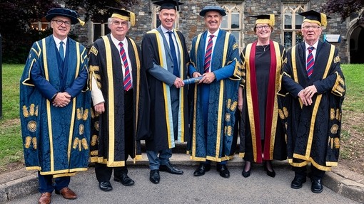 Ian Gwyn Hughes a’r Athro Dylan Jones yn gafael mewn sgrôl las; mae uwch aelodau eraill y Brifysgol yn sefyll ar y naill ochr iddynt; mae pawb mewn gwisg academaidd.