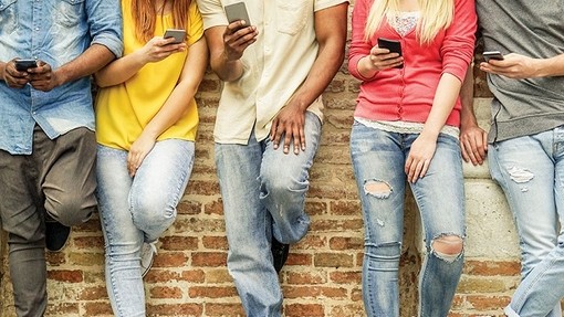 Five young people in ripped jeans leaning against a brick wall and holding their phones.